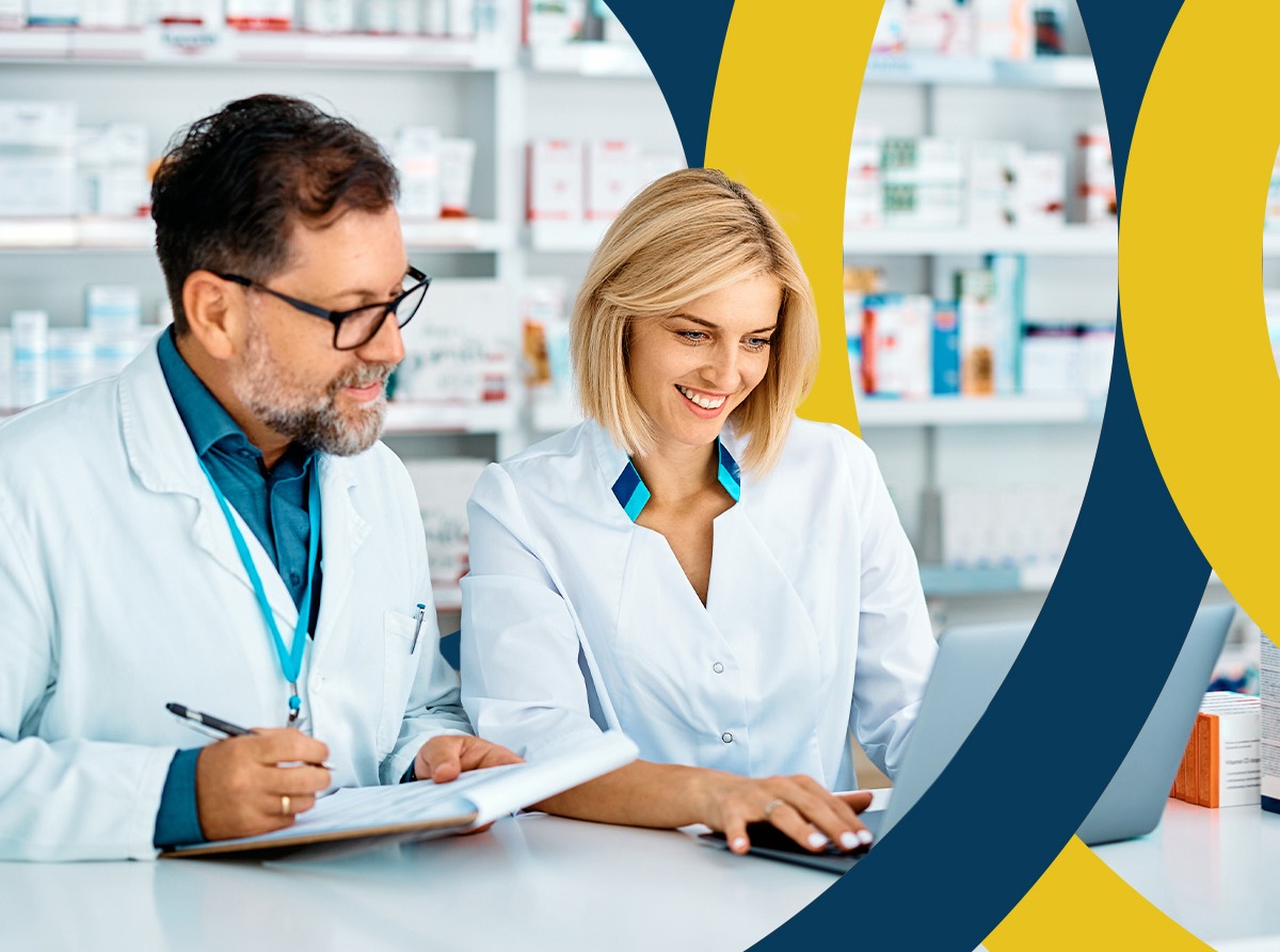 Pharmacist writing on paper records while a colleague checks the same work on a laptop at the pharmacy counter